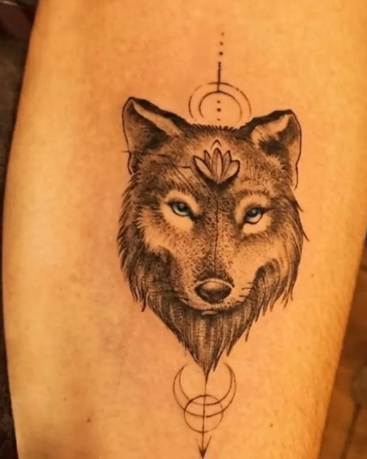 Tatuagem de lobo com olhos azuis, flor de lótus na testa e luas geométricas no antebraço