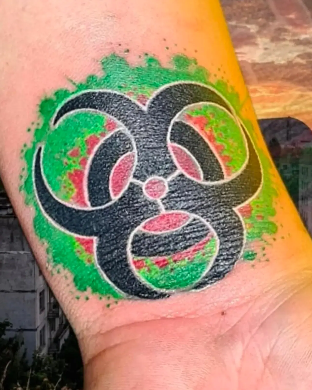 Tatuagem colorida no pulso: símbolo de risco biológico preto sobre mancha verde e vermelha
