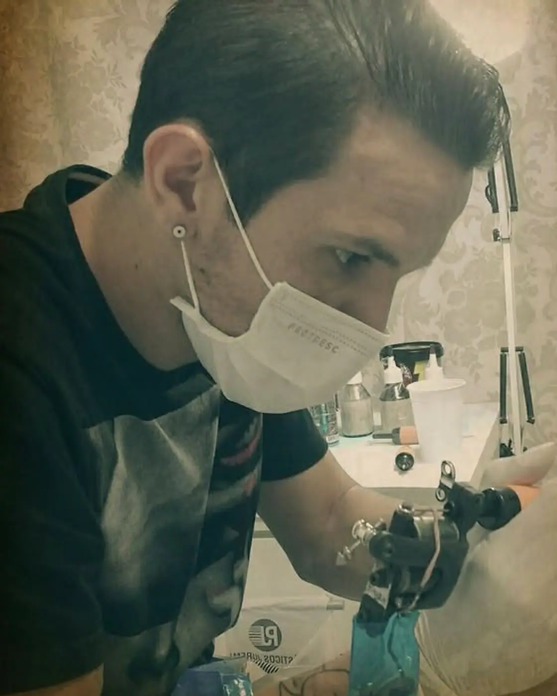 Manshah Marcelo tatuando no estúdio, de máscara, com a máquina na mão